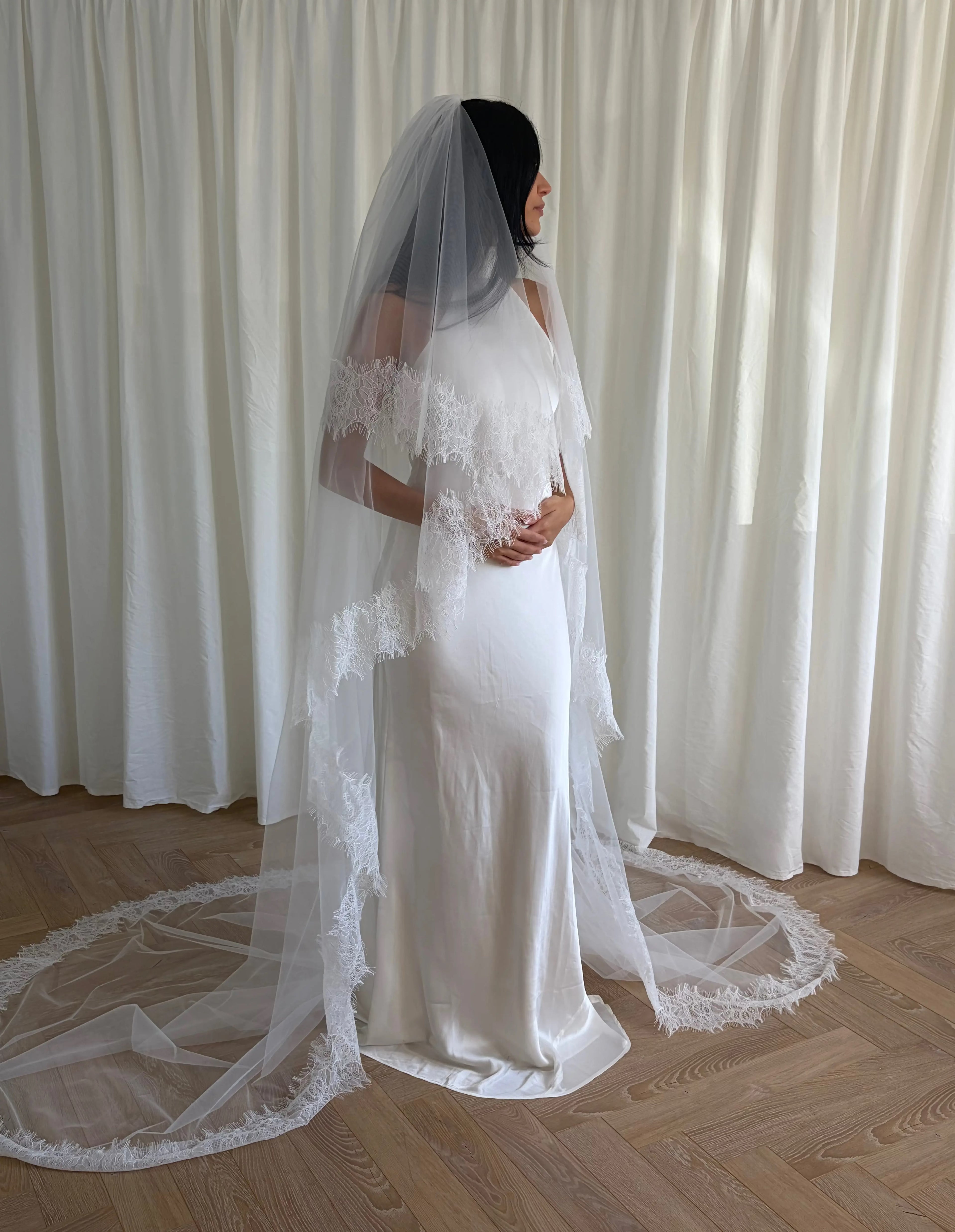 This cathedral-length lace trim blusher wedding veil flows gracefully behind the bride, made from soft, lightweight tulle and finished with intricate lace trim. Ideal for traditional or formal weddings, it creates a striking, romantic silhouette and complements any bridal gown with elegance and sophistication.