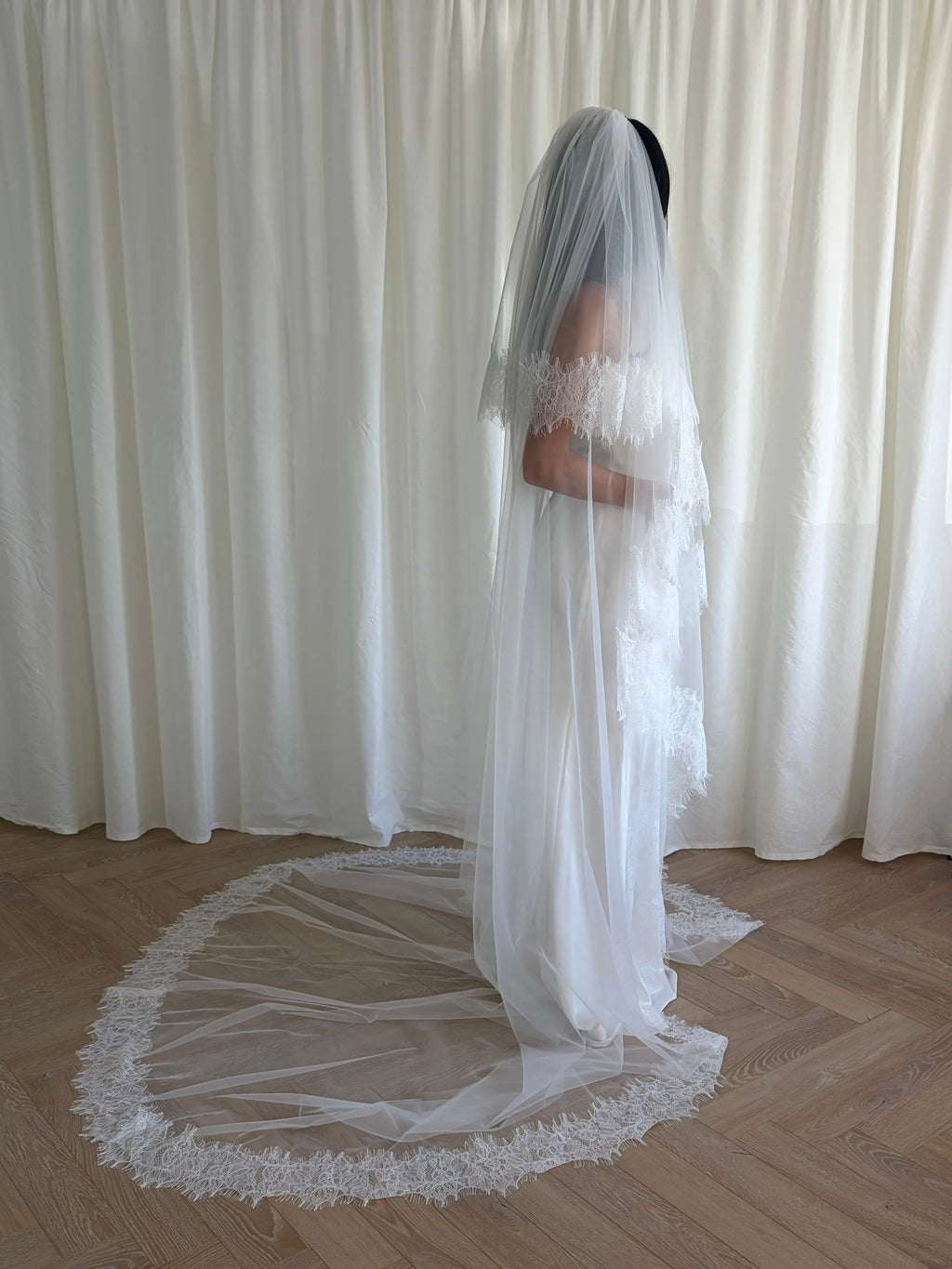 This cathedral-length lace trim blusher wedding veil flows gracefully behind the bride, made from soft, lightweight tulle and finished with intricate lace trim. Ideal for traditional or formal weddings, it creates a striking, romantic silhouette and complements any bridal gown with elegance and sophistication.
