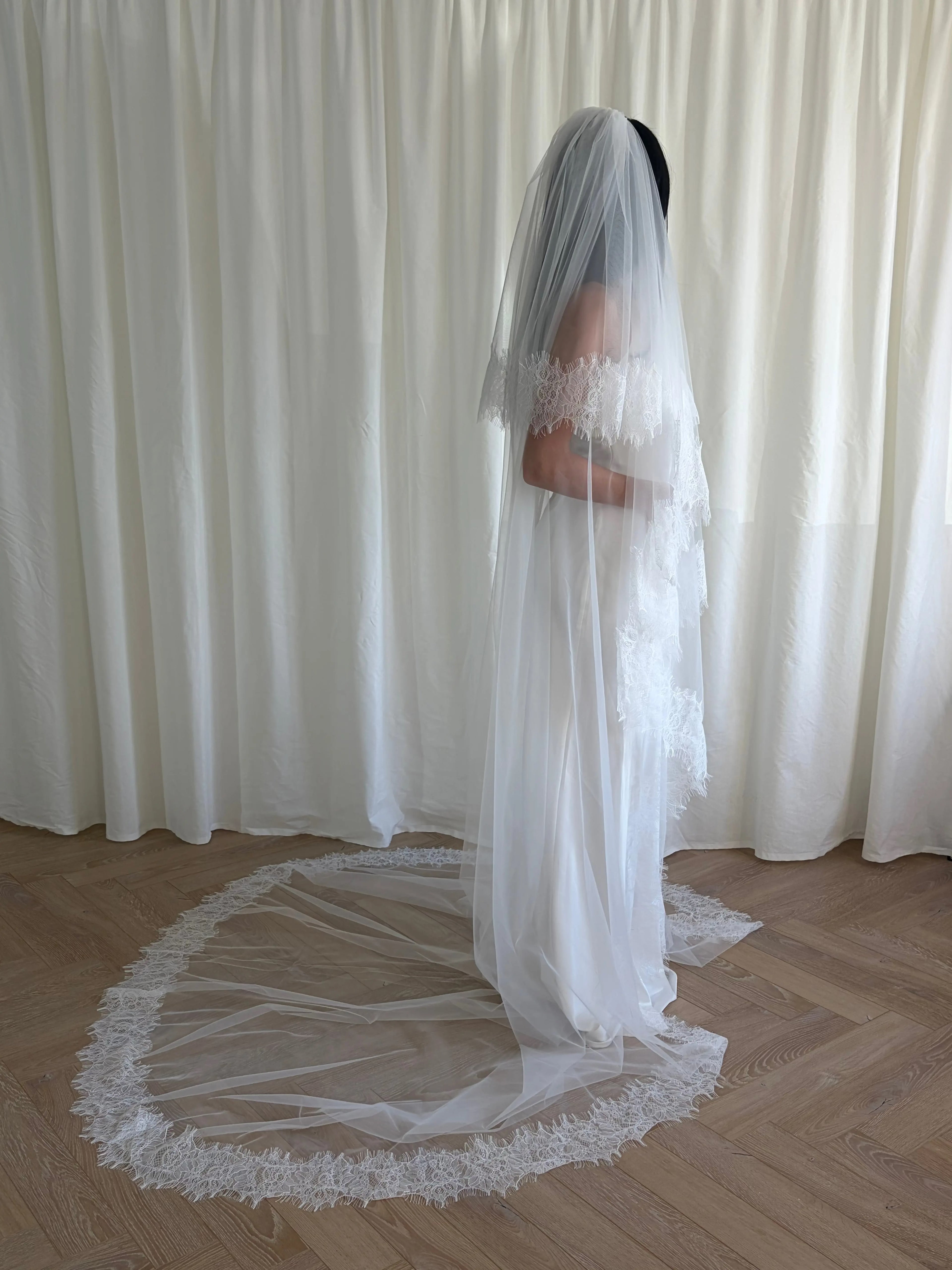 This cathedral-length lace trim blusher wedding veil flows gracefully behind the bride, made from soft, lightweight tulle and finished with intricate lace trim. Ideal for traditional or formal weddings, it creates a striking, romantic silhouette and complements any bridal gown with elegance and sophistication.