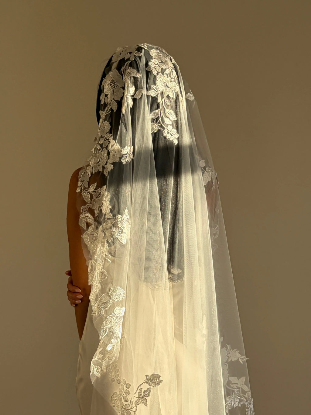 The Embroidered Ethereal Garden Floral Lace Veil features fine sequin embroidery and hand-finished floral lace trim for a soft, ethereal glow. Crafted from luxurious tulle in a soft off-white tone, this romantic bridal veil drapes beautifully for a fairytale-inspired finish. Perfect for garden or cathedral weddings.