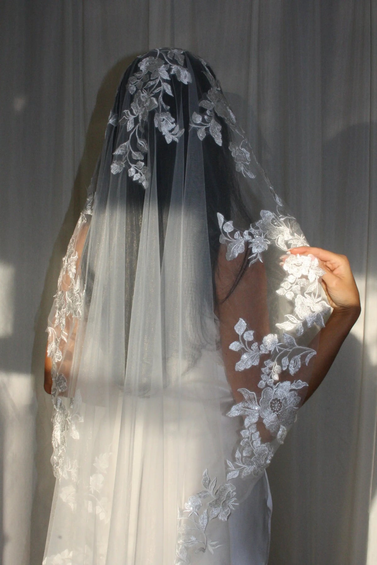 The Embroidered Ethereal Garden Floral Lace Veil features fine sequin embroidery and hand-finished floral lace trim for a soft, ethereal glow. Crafted from luxurious tulle in a soft off-white tone, this romantic bridal veil drapes beautifully for a fairytale-inspired finish. Perfect for garden or cathedral weddings.