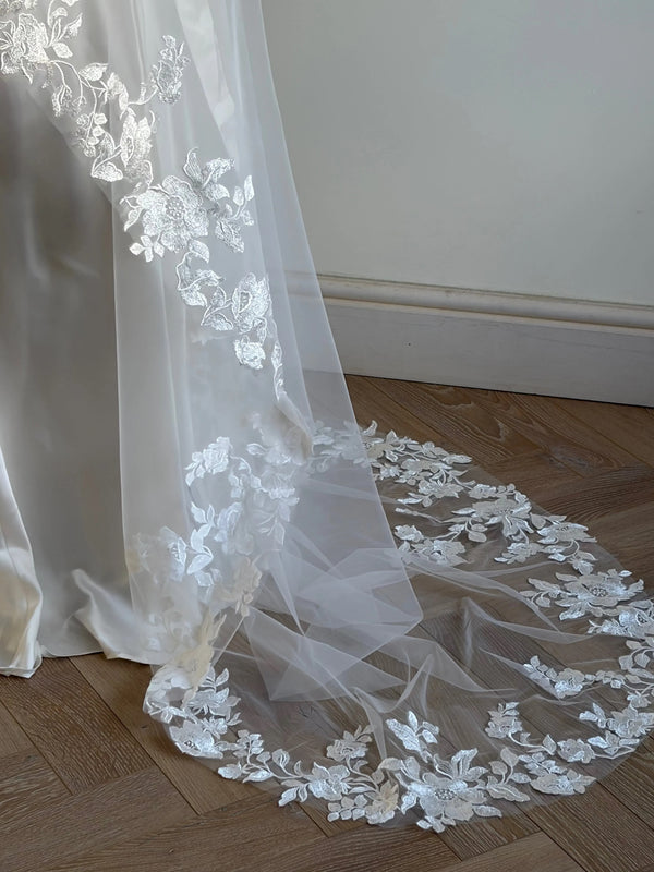 The Embroidered Ethereal Garden Floral Lace Veil features fine sequin embroidery and hand-finished floral lace trim for a soft, ethereal glow. Crafted from luxurious tulle in a soft off-white tone, this romantic bridal veil drapes beautifully for a fairytale-inspired finish. Perfect for garden or cathedral weddings.
