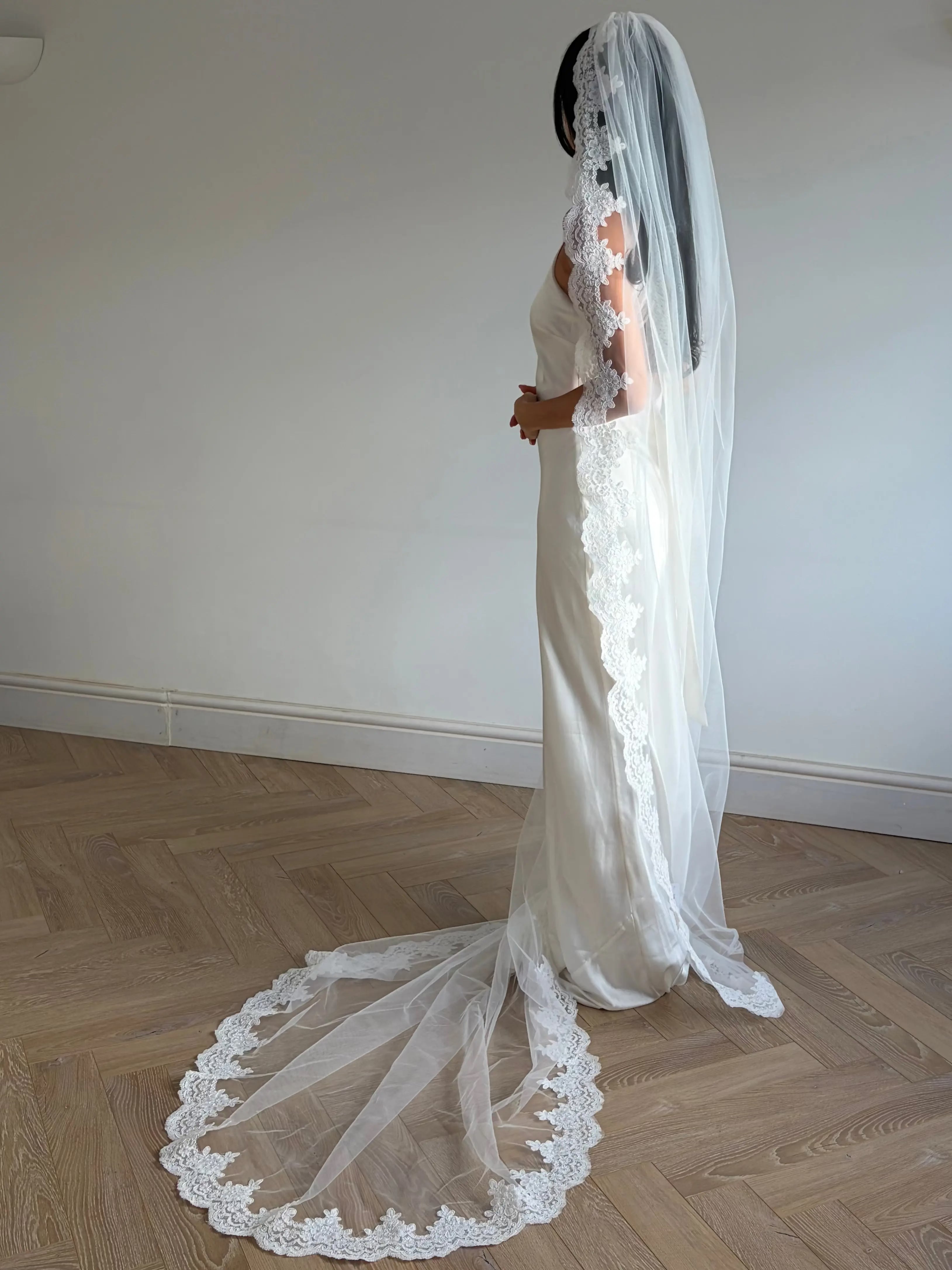 This pre-order lace trim bunched mantilla veil combines cathedral-length drama with hauntingly soft elegance. Made from luxurious, lightweight tulle and finished with fine lace edging, it falls straight to the ground in a flowing, cloak-like drape. Designed for brides seeking a romantic and timeless silhouette