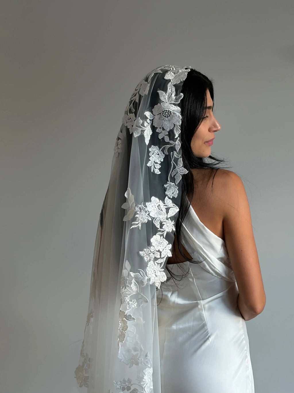 The Embroidered Ethereal Garden Floral Lace Veil features fine sequin embroidery and hand-finished floral lace trim for a soft, ethereal glow. Crafted from luxurious tulle in a soft off-white tone, this romantic bridal veil drapes beautifully for a fairytale-inspired finish. Perfect for garden or cathedral weddings.