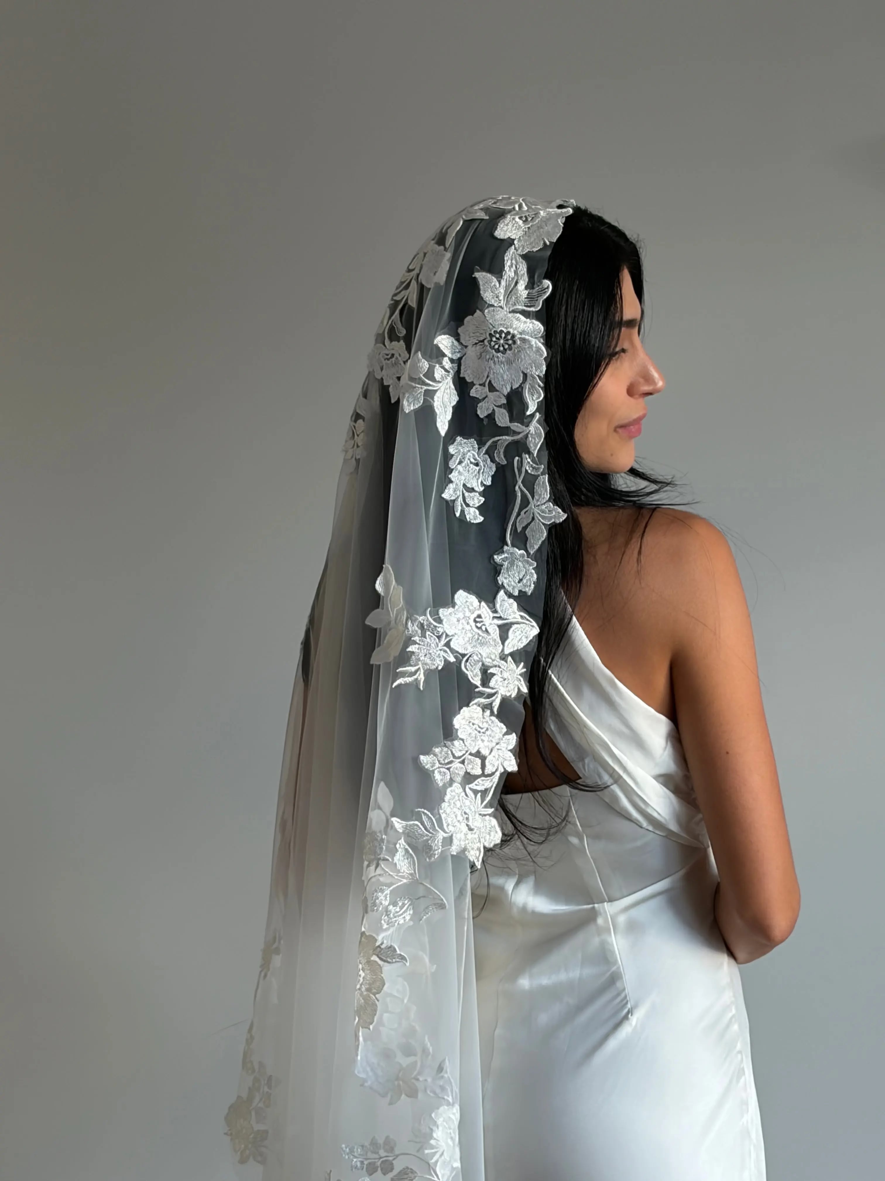 The Embroidered Ethereal Garden Floral Lace Veil features fine sequin embroidery and hand-finished floral lace trim for a soft, ethereal glow. Crafted from luxurious tulle in a soft off-white tone, this romantic bridal veil drapes beautifully for a fairytale-inspired finish. Perfect for garden or cathedral weddings.