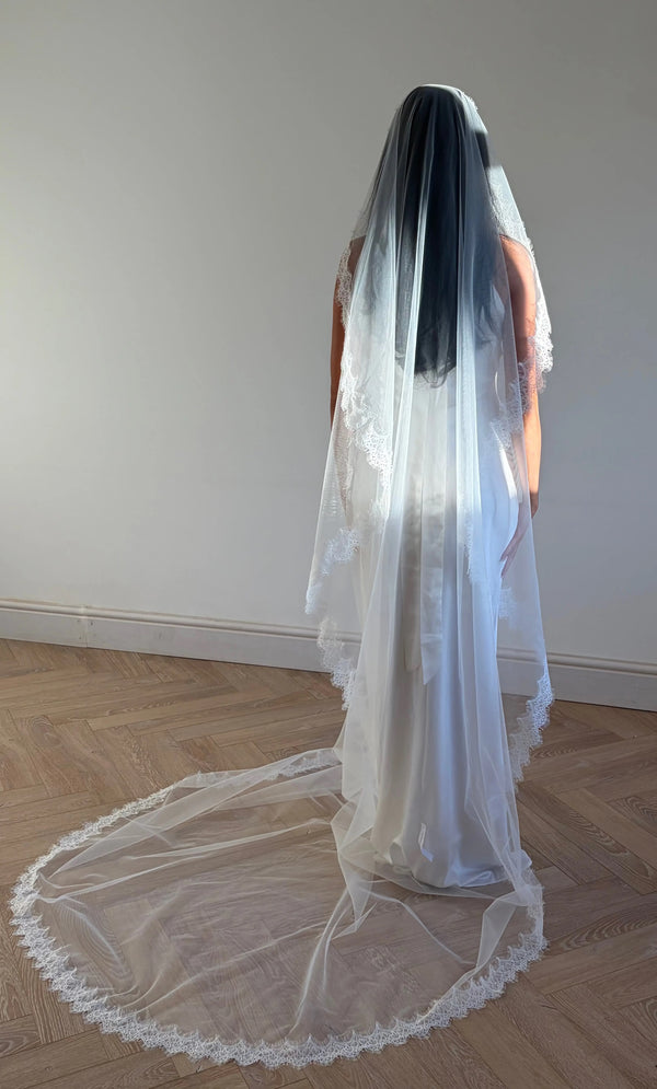 The Mantilla French Chantilly Lace Cathedral-Length Veil captures the romance of classic bridal style with exquisite craftsmanship and detail. Made from fine, lightweight tulle, this single-tier veil is bordered with intricate French Chantilly floral lace, complete with delicate eyelash edging that adds soft texture and movement. Its cathedral length creates a graceful, dramatic silhouette perfect for traditional or contemporary gowns. A hidden comb ensures a secure, comfortable hold, letting the veil flow 