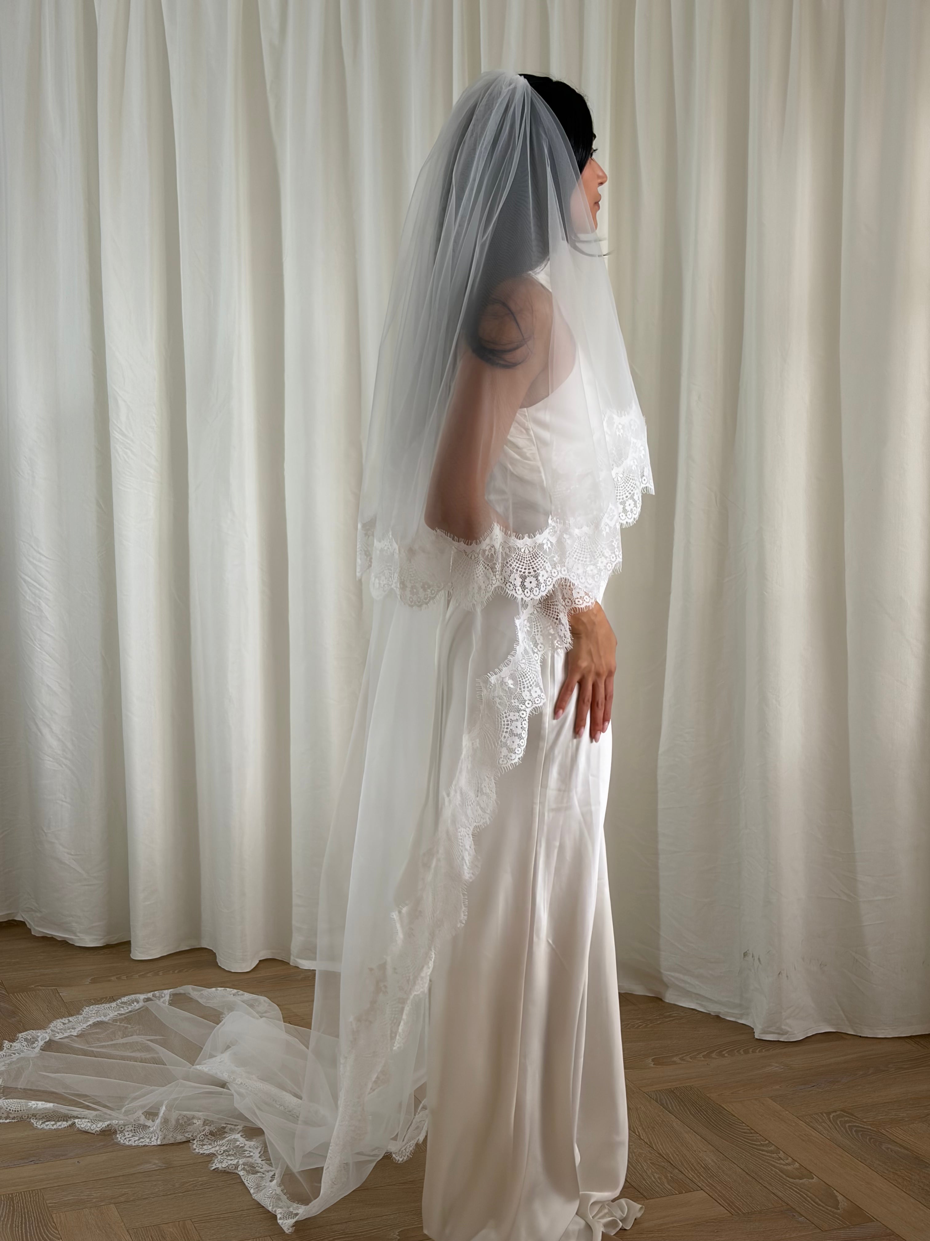 The Eyelash Trim Blusher Veil is a light, romantic accessory for modern brides seeking an ethereal finishing touch. Made from soft, flowing tulle, it features delicate eyelash lace along the edge for a subtle, feminine detail. The veil drapes softly over the face and shoulders, enhancing the bride’s silhouette with an airy, graceful effect. Its discreet comb attachment ensures a secure, seamless fit, making it ideal for both ceremony and photography. Perfect for adding a classic yet dreamy accent to any gow